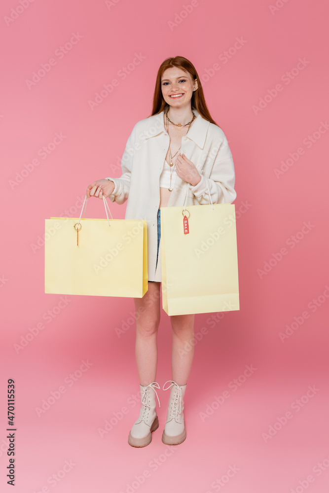 Full length of smiling red haired woman holding shopping bags with price tag and looking at camera on pink background