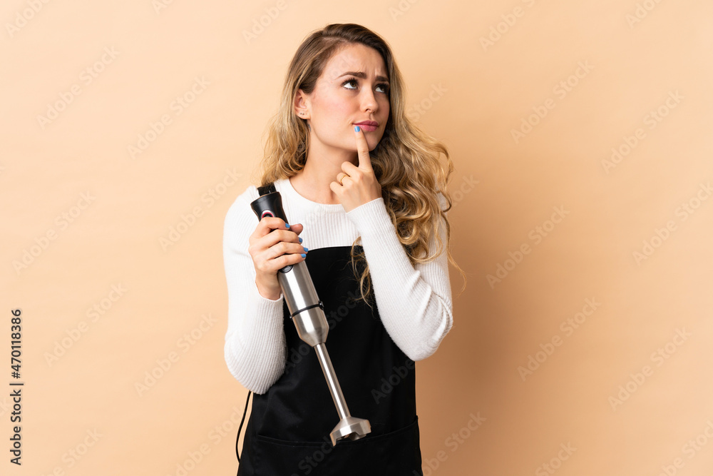 Young brazilian woman using hand blender isolated on beige background having doubts while looking up Young brazilian woman using hand blender isolated on beige background having doubts while looking up