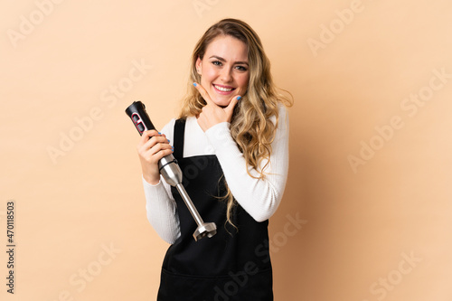 Young brazilian woman using hand blender isolated on beige background happy and smiling