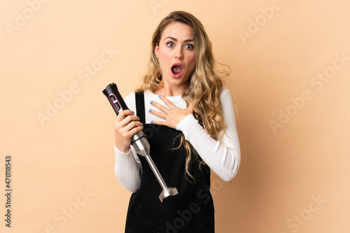 Young brazilian woman using hand blender isolated on beige background surprised and shocked while looking right