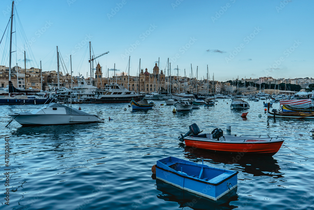 Traditional colorful Malta boats and luxury yachts in Birgu harbor at sunset,old town in background.Travel vacation concept.Boat trip in Mediterranean.View of expensive sailing yachts at pier