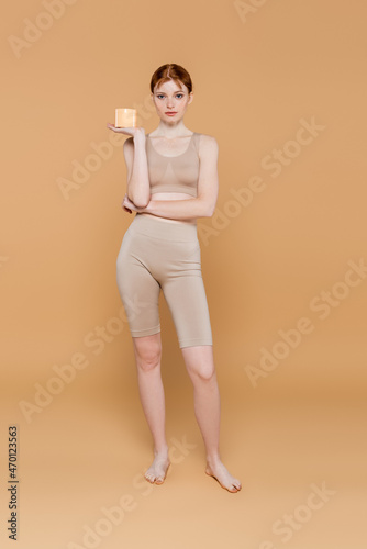 Full length of pretty redhead woman holding container with cosmetic cream on beige background © LIGHTFIELD STUDIOS