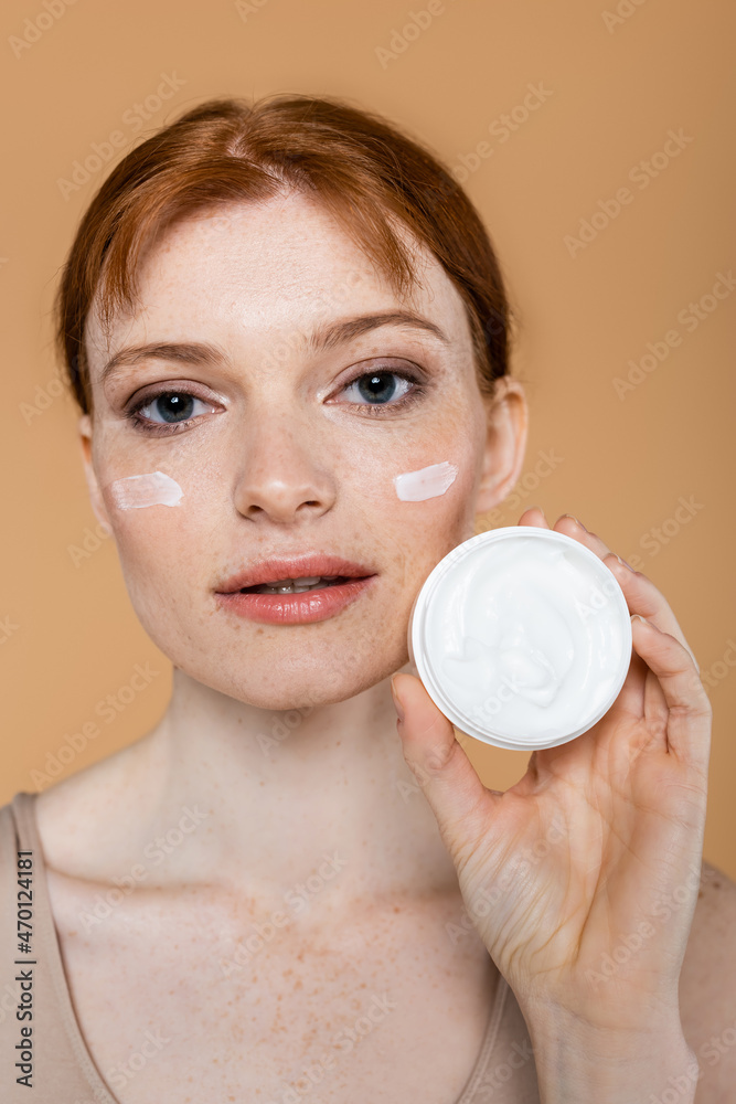 Young redhead woman showing container with cosmetic cream isolated on beige Young redhead woman showing container with cosmetic cream isolated on beige