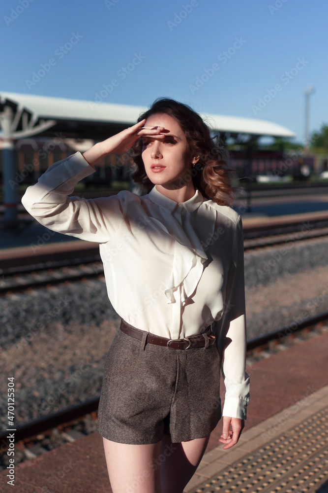 A young beautiful woman in a white shirt stands on the platform of the railway station, looks into the distance seeing off or meeting. The woman is waiting.