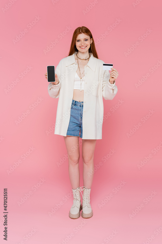 Pretty and stylish red haired woman holding cellphone and credit card on pink background Pretty and stylish red haired woman holding cellphone and credit card on pink background