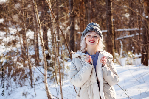 Smiling blonde woman in warm knitted cap and beige coat outdoors in winter forest