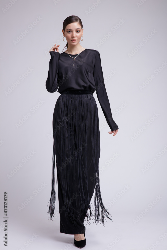 Fototapeta premium High fashion photo of a beautiful elegant young woman in a pretty black blouse, fringe trousers, accessories over white background. Studio Shot. Gathered dark hair. Slim figure
