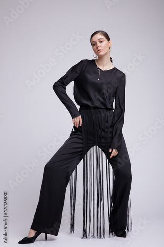 High fashion photo of a beautiful elegant young woman in a pretty black blouse, fringe trousers, accessories over white background. Studio Shot. Gathered dark hair. Slim figure