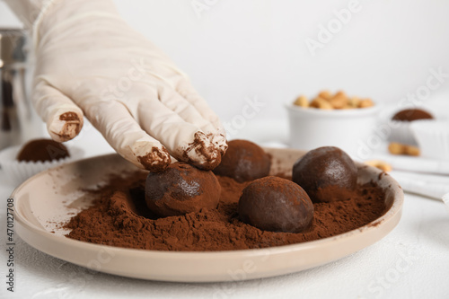 Confectioner covering delicious chocolate truffles with cocoa powder at white table, closeup
