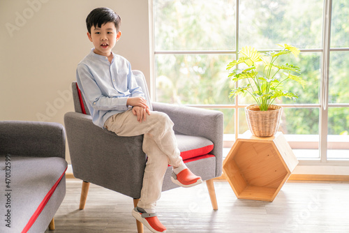 Asia boy wearing blue T-shirt, cross legged and sitting on the sofa in the living room with copy space. Background is the windows and sunlight. Plant pot beside kid. Lifestyle and relax