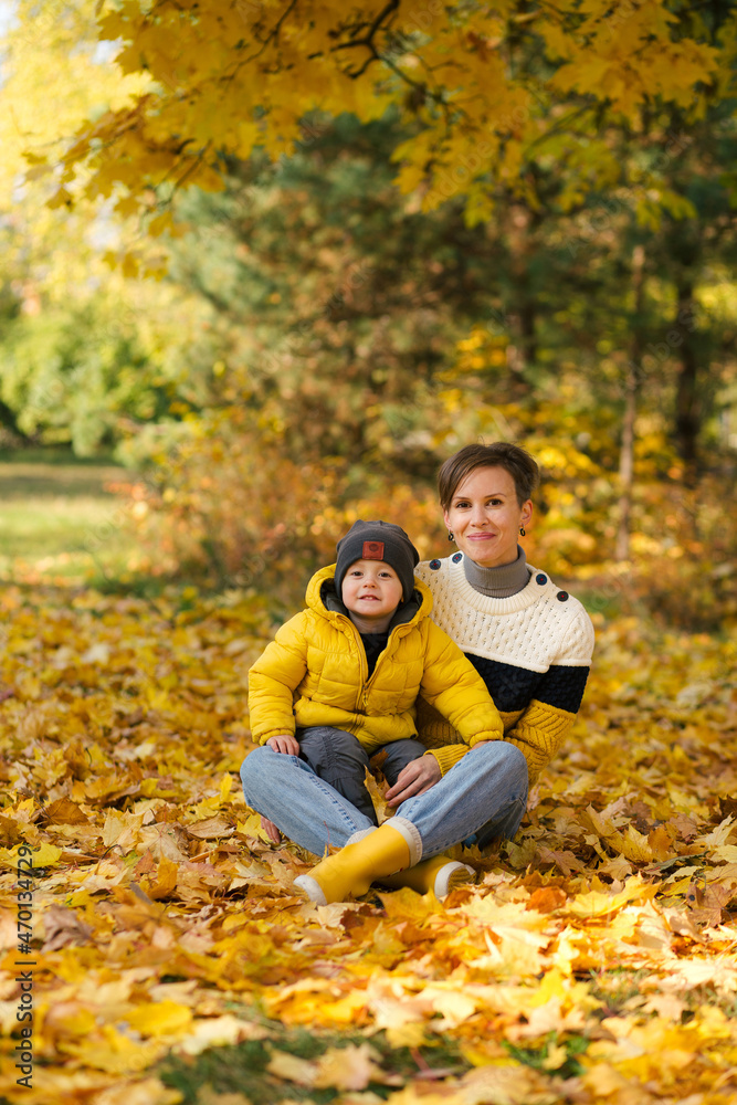 Happy young mother and little son sitting in autumn park among the yellow maple leaves