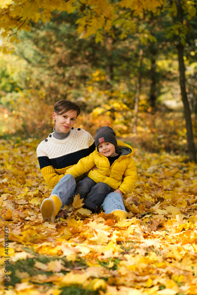 Happy young mother and little son sitting in autumn park among the yellow maple leaves Happy young mother and little son sitting in autumn park among the yellow maple leaves