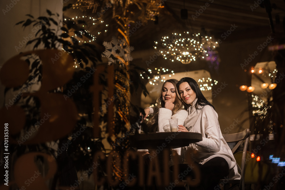 two beautiful young women drink coffee in a coffee shop in winter in a romantic setting. view through the window. blurry lights