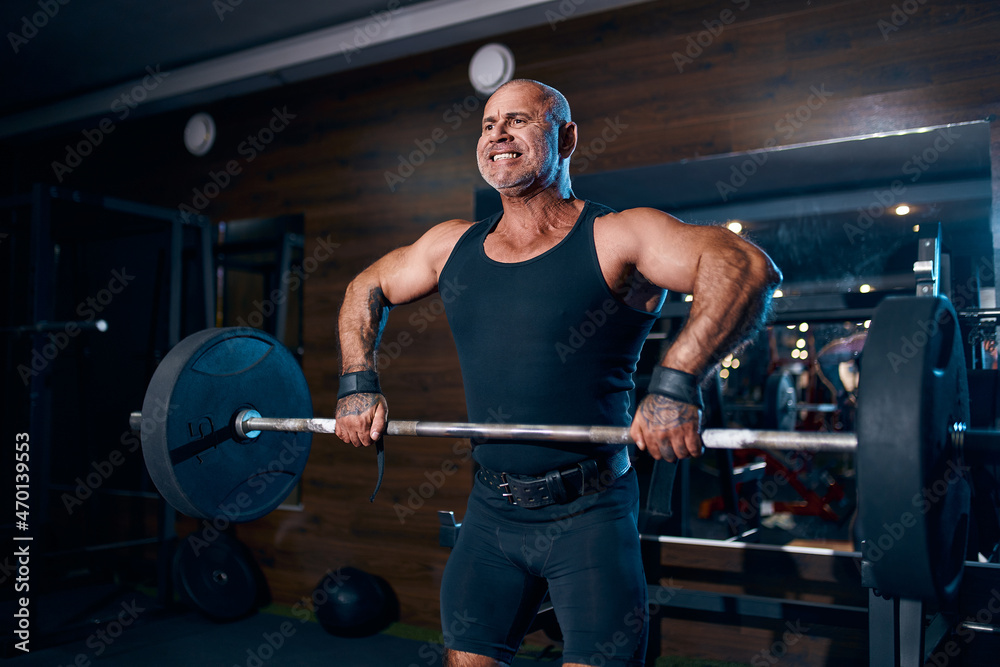 Emotional aged bald bodybuilder doing trapezius and deltoid exercise with a barbell in the gym