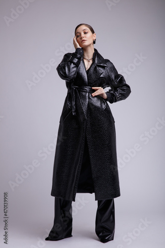 High fashion photo of a beautiful elegant young woman in a pretty leather coat, pants, accessories posing over white, soft gray background. Studio Shot. Gathered dark hair. Slim figure