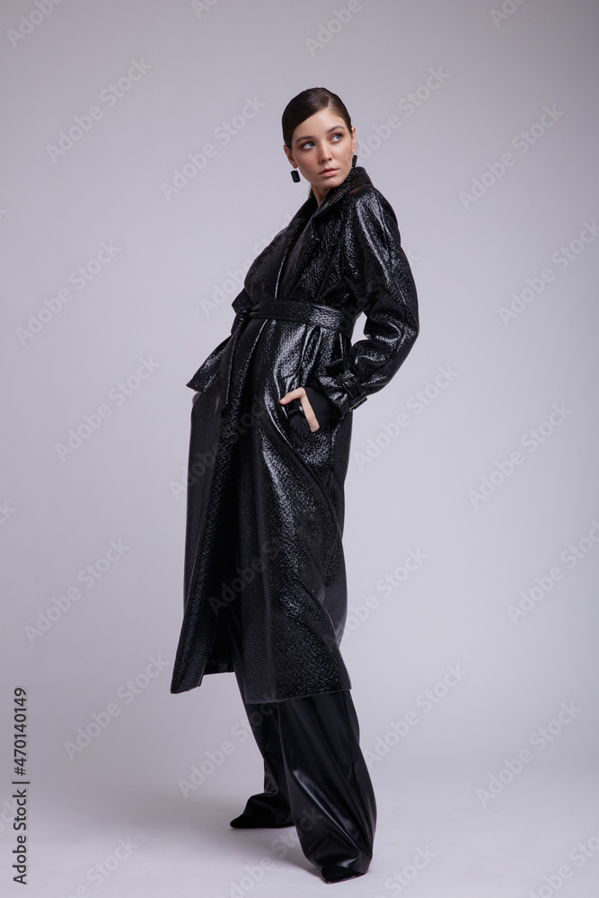 High fashion photo of a beautiful elegant young woman in a pretty leather coat, pants,  accessories posing over white, soft gray background. Studio Shot. Gathered dark hair. Slim figure
