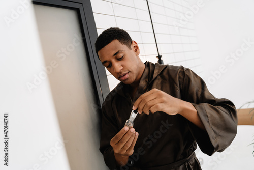 Black man wearing bathrobe applying face serum in bathroom