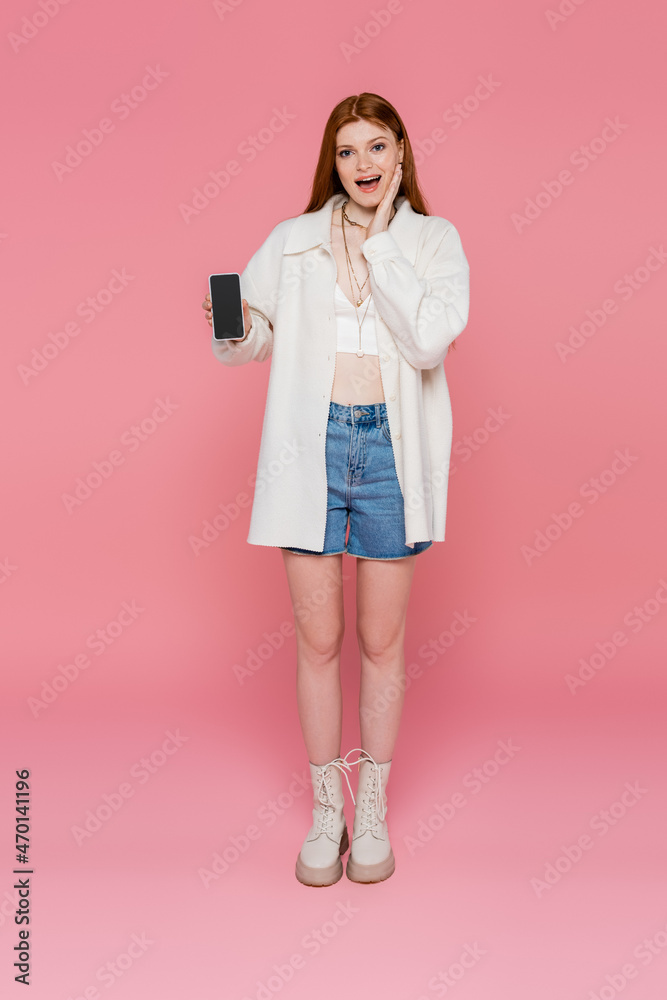 Excited woman in jacket and necklaces holding cellphone with blank screen on pink background Excited woman in jacket and necklaces holding cellphone with blank screen on pink background