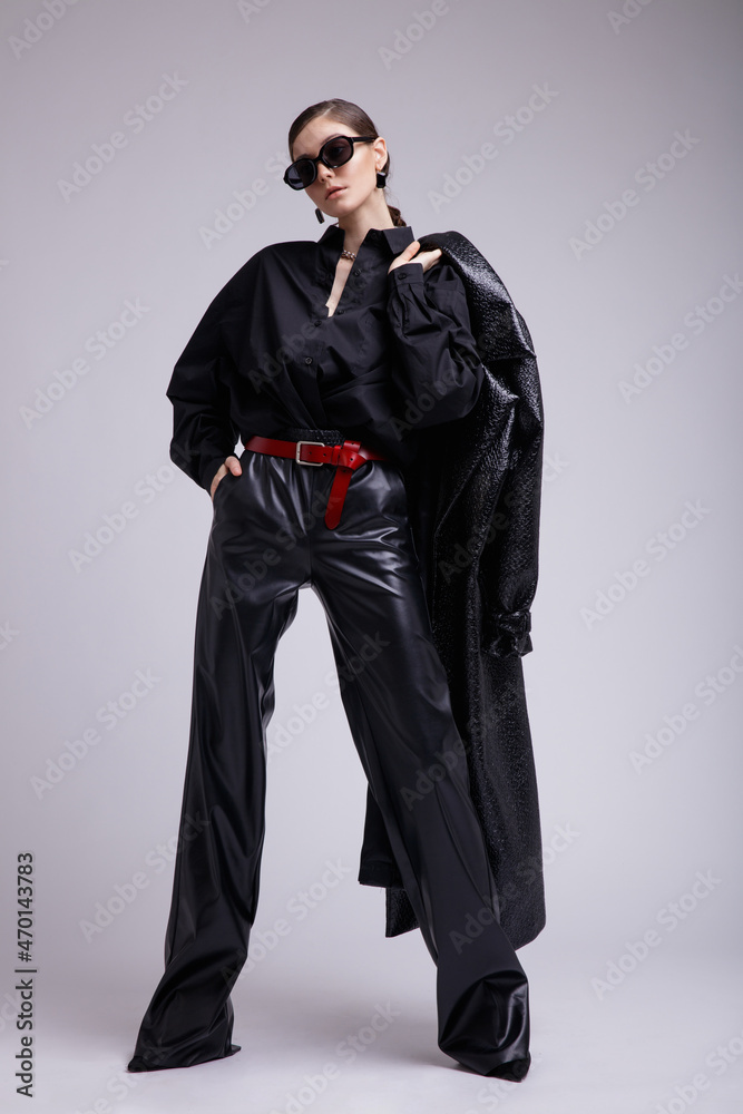 High fashion photo of a beautiful elegant young woman in a pretty leather coat, pants, stylish sunglasses, accessories, shirt, red belt posing over gray background. Studio Shot. Gathered dark hair High fashion photo of a beautiful elegant young woman in a pretty leather coat, pants, stylish sunglasses, accessories, shirt, red belt posing over gray background. Studio Shot. Gathered dark hair