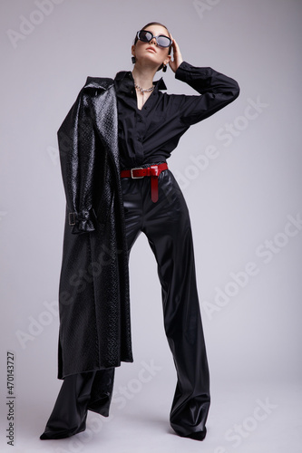 High fashion photo of a beautiful elegant young woman in a pretty leather coat, pants, stylish sunglasses, accessories, shirt, red belt posing over gray background. Studio Shot. Gathered dark hair