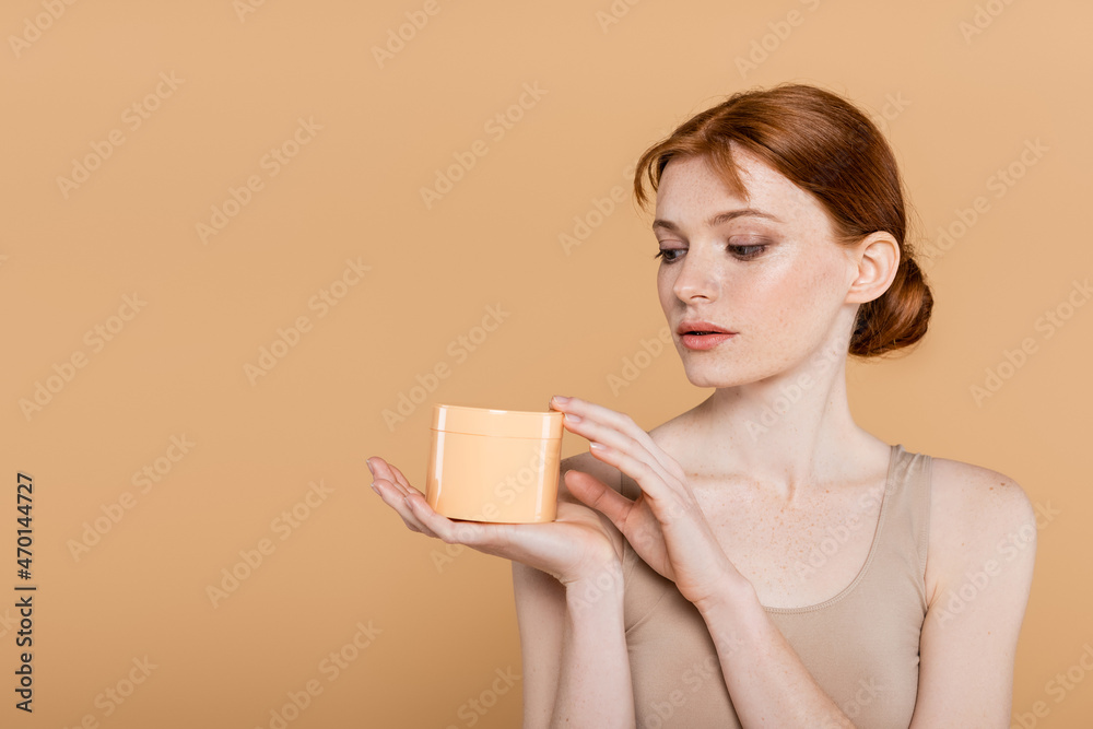 Redhead woman with freckles holding cosmetic cream isolated on beige