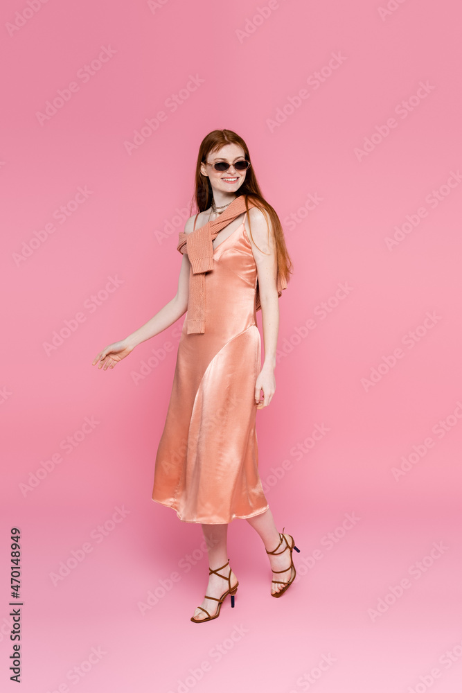 Stylish woman in silk dress and sunglasses walking on pink background