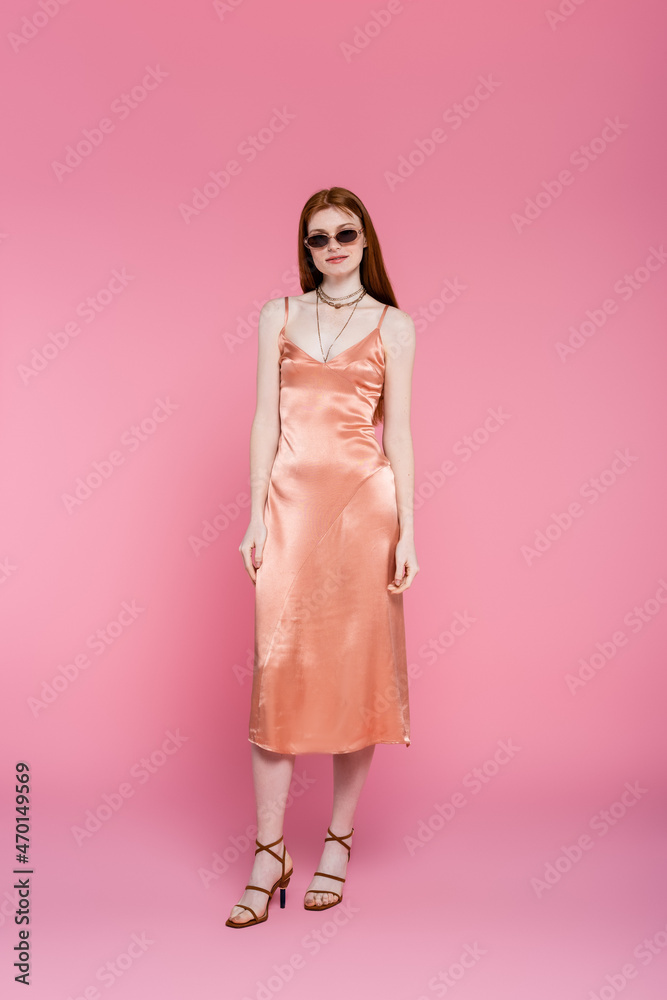 Full length of young red haired woman in sunglasses and dress standing on pink background Full length of young red haired woman in sunglasses and dress standing on pink background