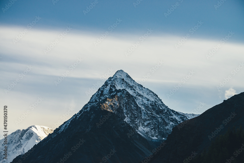 Sunny landscape with great mountains silhouettes and snow-covered pointy peak with golden sunshine on rocks. Mountain with peaked top with snow in sunshine. Beautiful pointed pinnacle in sunlight.