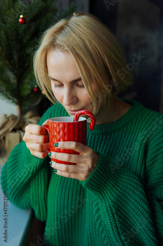 A lovely woman in a green sweater is holding a red cup with a hot drink in her hands, drinking coffee or tea against the background of a Festive Christmas Tree. The holiday of Christmas and New Year.