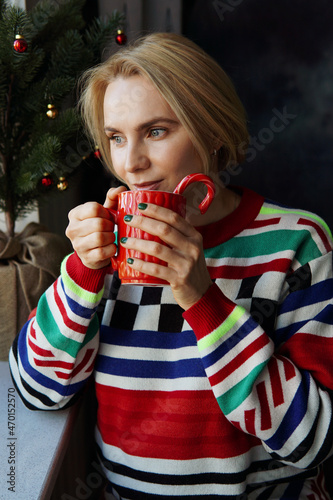 A lovely woman in a bright sweater is holding a red cup with a hot drink in her hands, drinking coffee or tea against the background of a Festive Christmas Tree. The holiday of Christmas and New Year.