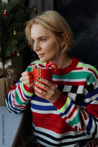 A lovely woman in a bright sweater holds a red cup with a hot drink in her hands and looks out the window against the background of a Festive Christmas Tree. The holiday of Christmas and New Year.