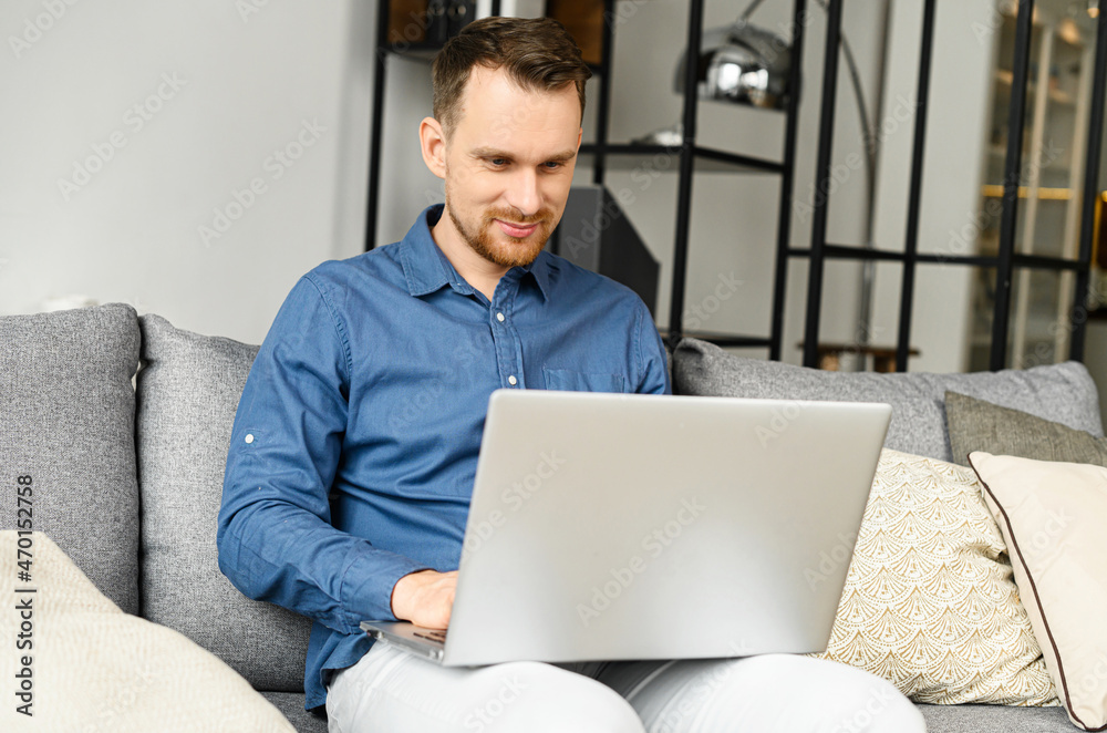 Smiling young bearded man is using a laptop at home, a guy looks at the device screen with interest sitting on the couch, websurfing, watching news feed, works remotely from home Smiling young bearded man is using a laptop at home, a guy looks at the device screen with interest sitting on the couch, websurfing, watching news feed, works remotely from home