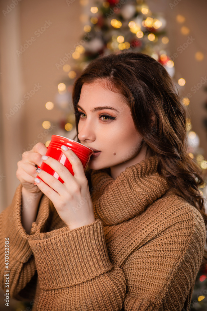 A beautiful girl in a warm sweater against the background of a Christmas tree and garlands is drinking coffee from a red mug. Model against the background of Christmas decorations. hot drink A beautiful girl in a warm sweater against the background of a Christmas tree and garlands is drinking coffee from a red mug. Model against the background of Christmas decorations. hot drink