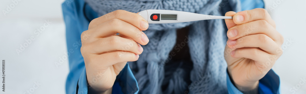 Fototapeta premium cropped view of sick woman in knitted scarf holding electronic thermometer, banner.