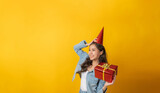 young pretty asian female in denim Jacket clothing smiling with red gift box board with copy space area for text on yellow background, holiday festival, christmas celebration, happy hew year concept