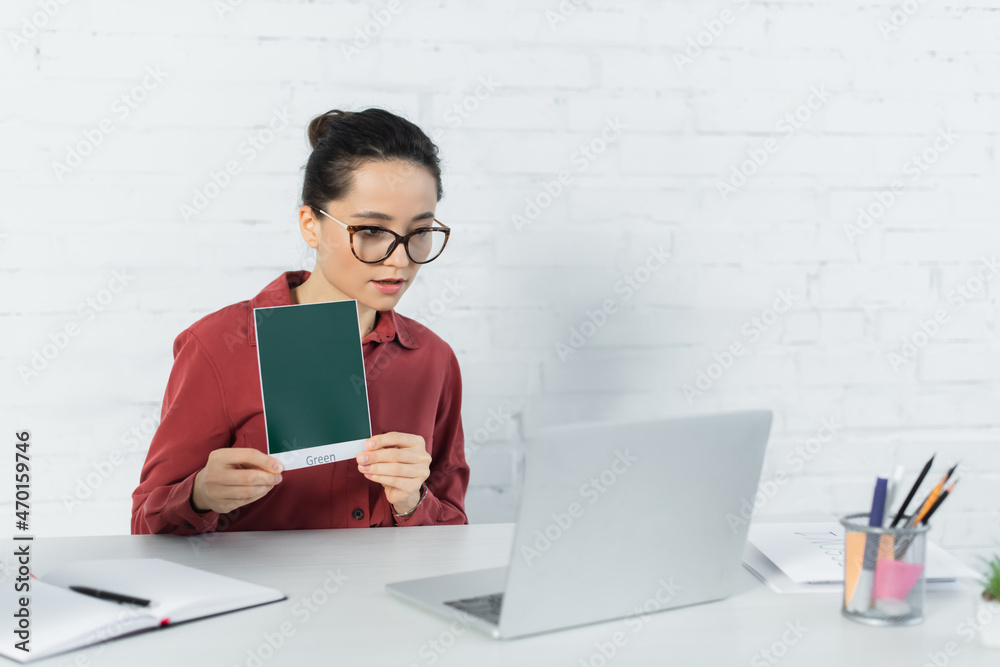 Obraz premium young teacher in eyeglasses holding card with green lettering and looking at laptop on desk.