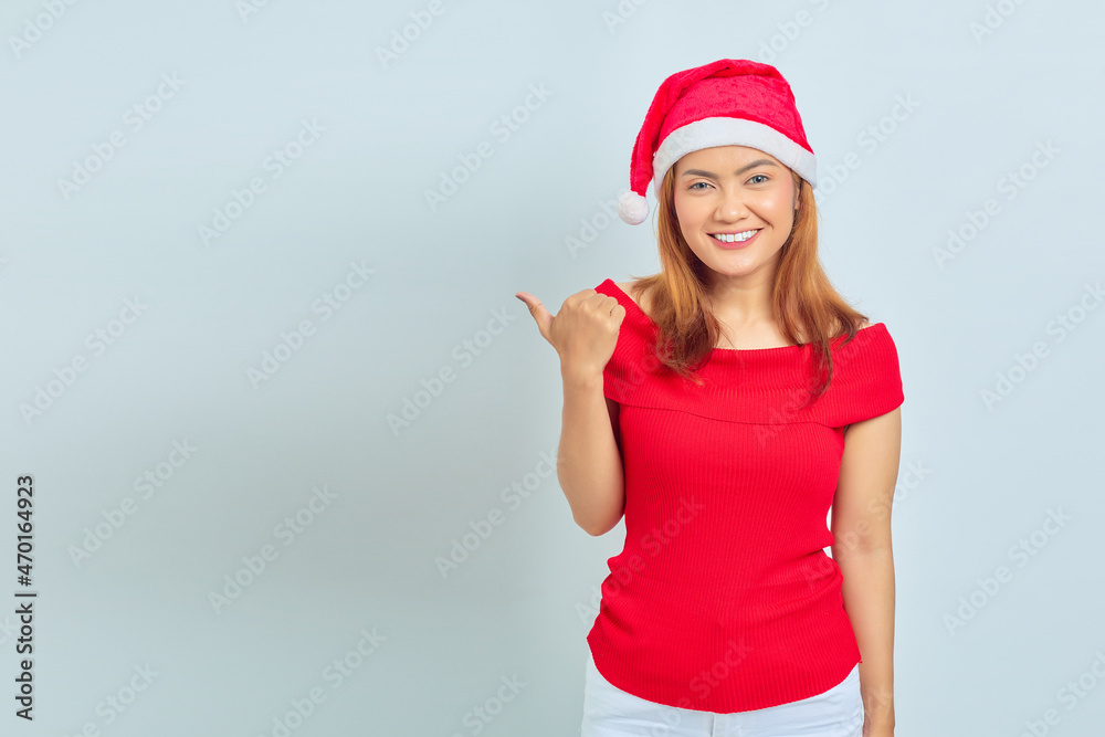 Photo of beautiful Asian girl in christmas hat with smiling facial expression and pointing aside on white background
