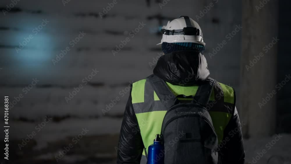 Adult Caucasian man with backpack in helmet with flashlight turning looking around at night outdoors. Portrait of confident tourist travelling on winter standing at building covered with snow