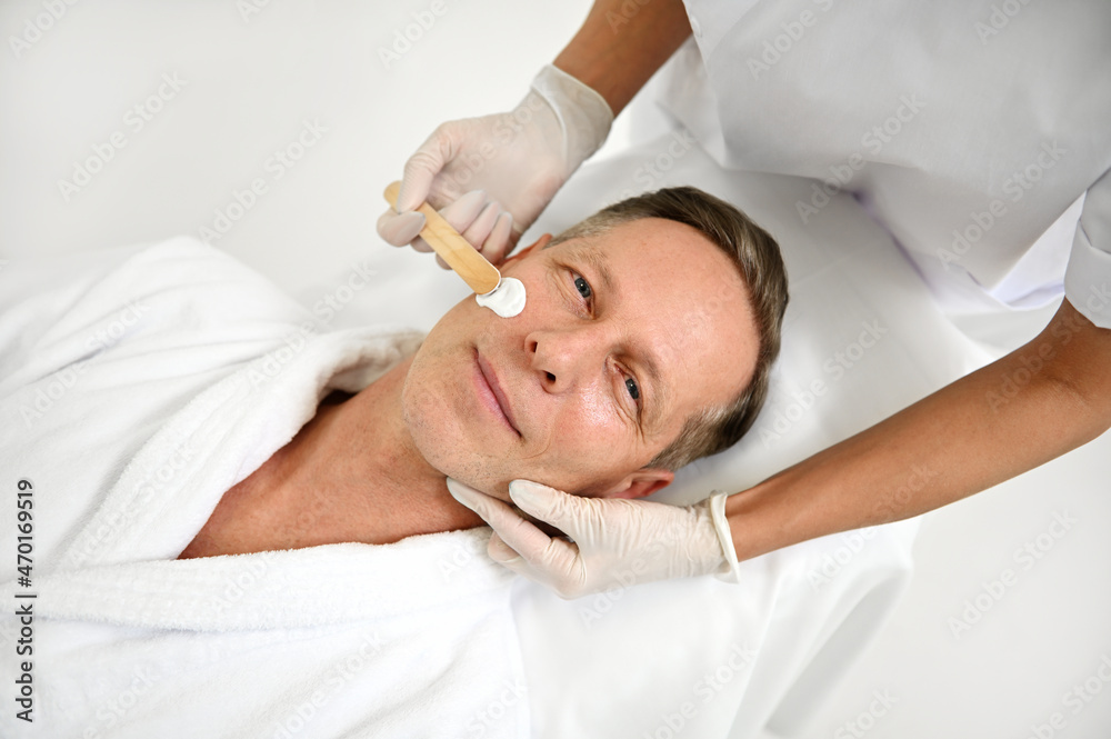 Top view of relaxed handsome middle aged man, lying on a massage table at wellness spa salon and looking at camera while beautician applying nourishing cosmetic product on his face. Male beauty care Top view of relaxed handsome middle aged man, lying on a massage table at wellness spa salon and looking at camera while beautician applying nourishing cosmetic product on his face. Male beauty care
