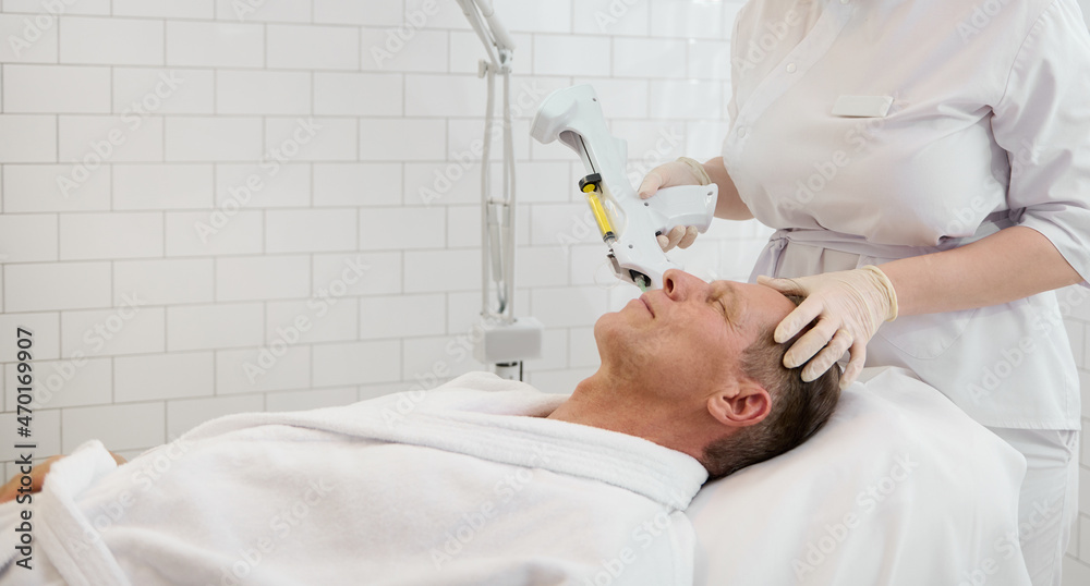 Handsome senior man wearing white terry robe, lying on massage table and receiving facial beauty injection with modern equipment in white luxury wellness spa. Anti-aging therapy, rejuvenation concept