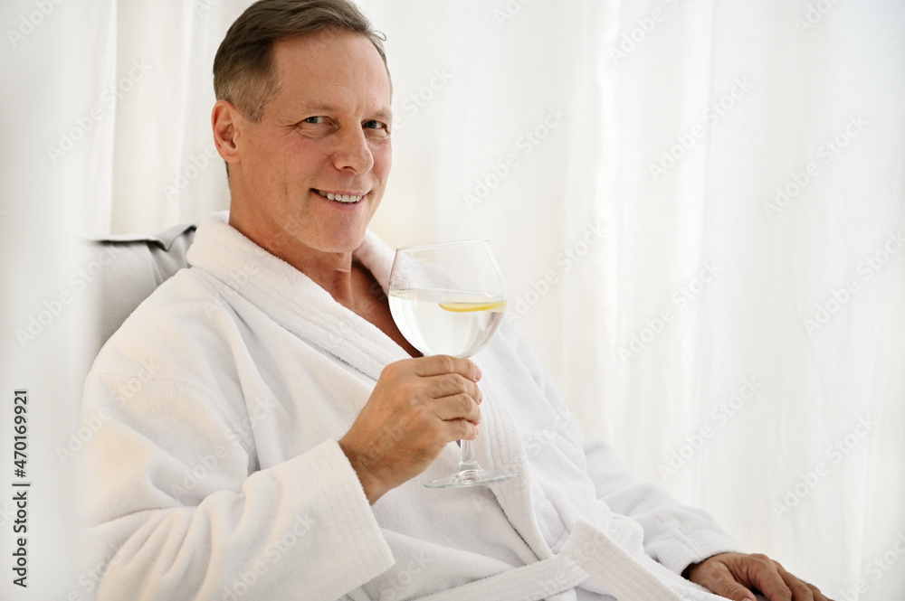 Handsome successful middle aged European man in white terry dressing gown holding glass of lemon water and smiling toothy smile looking at camera while relaxing in private room of wellness spa Handsome successful middle aged European man in white terry dressing gown holding glass of lemon water and smiling toothy smile looking at camera while relaxing in private room of wellness spa