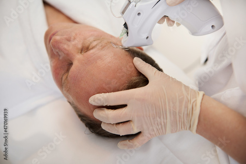 Overhead view of a handsome aged man lying on massage table during mesotherapy gun therapy in modern spa clinic. Anti-aging, rejuvenation and alternative beauty treatment to prevent first wrinkles