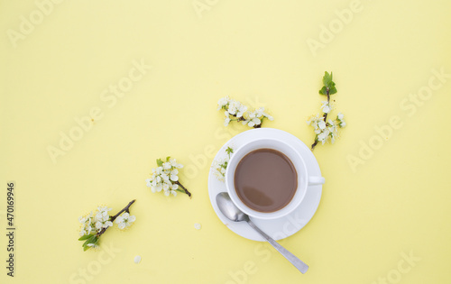 a white cup with cocoa on a yellow background, a teaspoon, branches of a flowering apple tree. Top view.