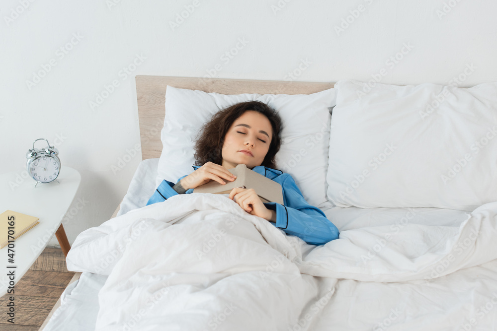 brunette woman in blue pajamas sleeping with book in bed.
