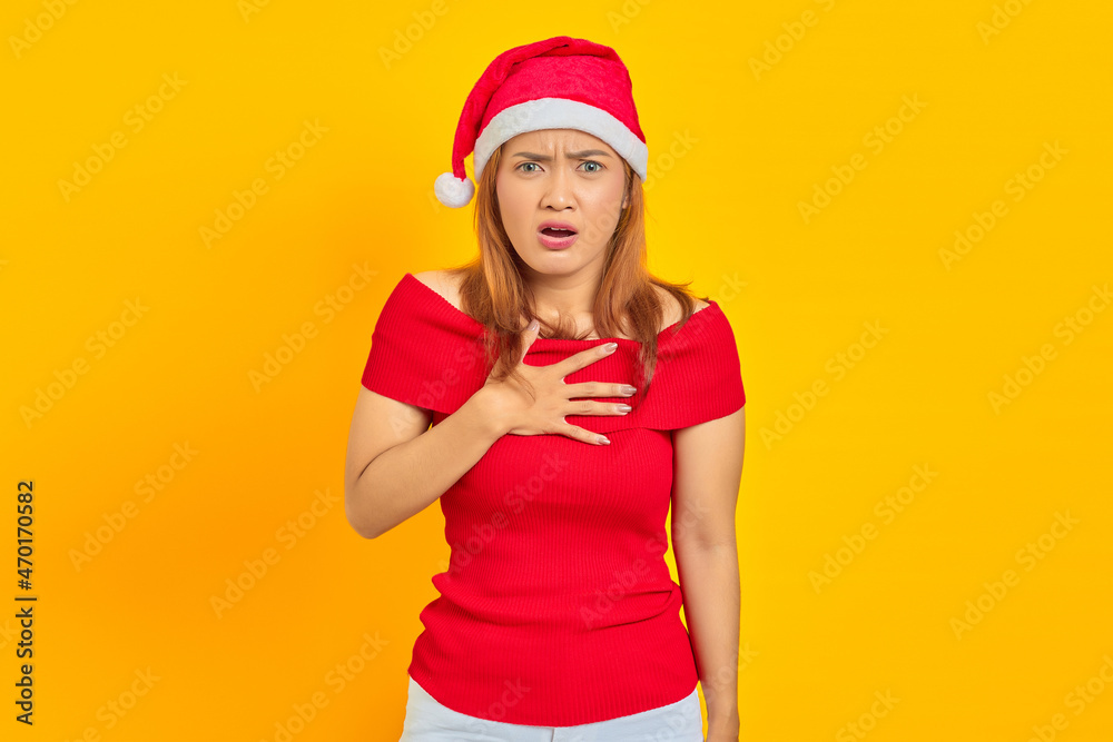 Shocked young Asian woman wearing Christmas hat and put hands chest on yellow background Shocked young Asian woman wearing Christmas hat and put hands chest on yellow background