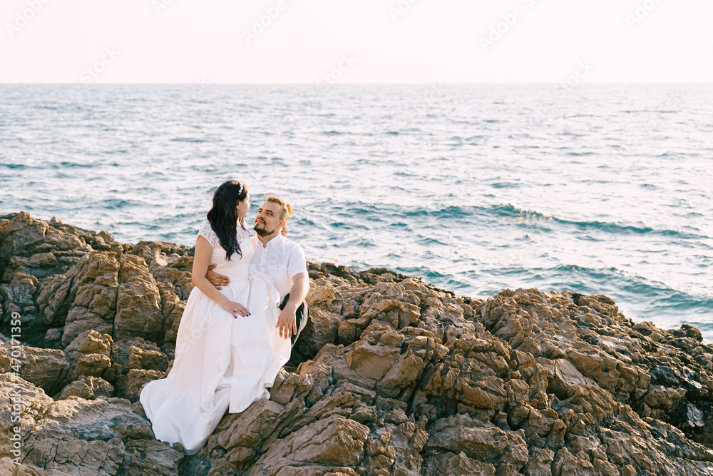 Bride sits on the knees of groom on the rocky seashore