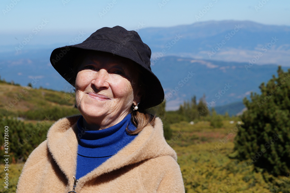 portrait of an elderly smiling woman in a black panama hat against the backdrop of a mountain landscape