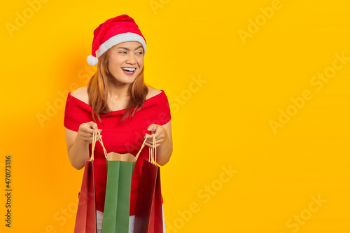 Cheerful young Asian woman in Santa Claus hat opening shopping bag and looking aside yellow background