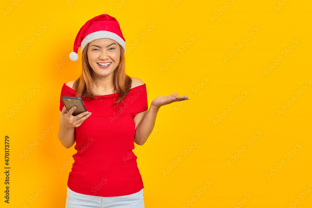 Portrait of smiling young Asian woman in Santa Claus hat using smartphone and showing copy space in palms over yellow background Portrait of smiling young Asian woman in Santa Claus hat using smartphone and showing copy space in palms over yellow background