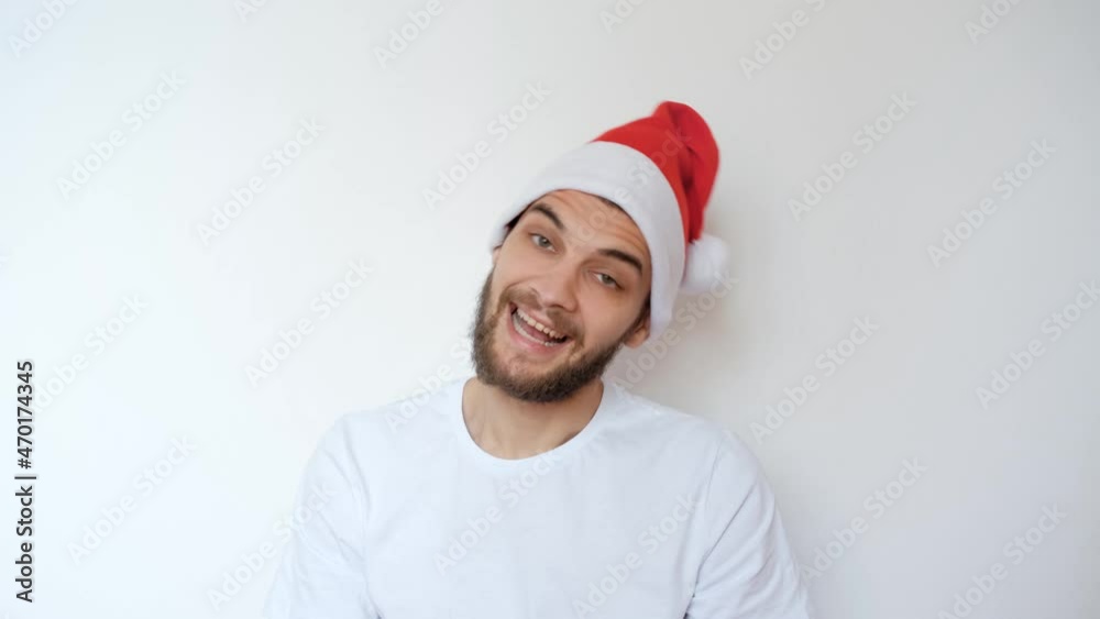 Caucasian handsome man in red Santa hat on white background smiles and waves his head in different directions. Christmas joy and emotions. Celebrate New year. Minimalism. 4K footage. Caucasian handsome man in red Santa hat on white background smiles and waves his head in different directions. Christmas joy and emotions. Celebrate New year. Minimalism. 4K footage.
